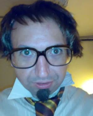 Colin Morgan with dark wig, large glasses, and striped tie - Comedic characterization.